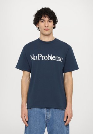 Mornarska pamučna majica s podebljim bijelim natpisom "No Problemo" u zaobljenom fontu. Kratki rukavi i okrugli izrez. Nosena s svjetloplavim trapericama.