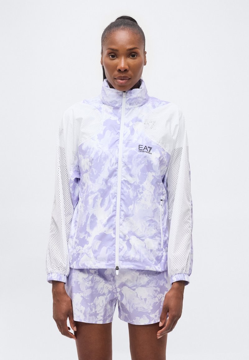 Γυναίκα φοράει ανοιχτό μοβ και λευκό ζακέτα με φερμουάρ και ταιριαστό σορτς, στέκεται μπροστά σε λευκό, απλό φόντο.