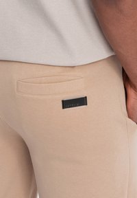 I pantaloni jogger beige presentano una texture liscia, una tasca posteriore e un'etichetta nera rettangolare con la scritta "ombre" in lettere bianche.