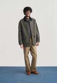 Χακί πανωφόρι από καμβά με σκούρο γιακά, με μπροστινές τσέπες, κουμπιά και κεντημένο λογότυπο. Συνδυάζεται με ανοιχτό καφέ παντελόνι και καφέ παπούτσια.