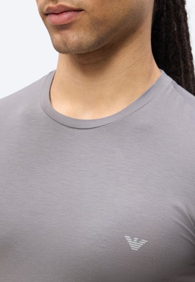 Κοντινό πλάνο ενός ατόμου που φοράει γκρι μπλούζα με στρογγυλή λαιμόκοψη και μικρό ριγέ λογότυπο στο στήθος, εμφανίζοντας μέρος του λαιμού και της γραμμής του σαγονιού.