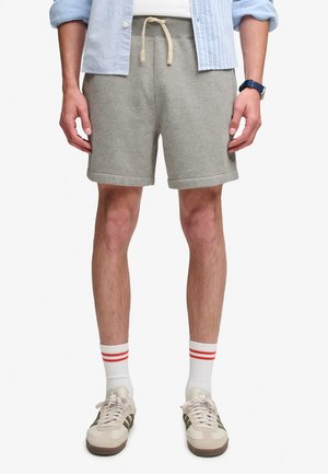 Mand iført lysegrå snøretrusser, hvide sneakers med grønne striber, hvide sokker med røde striber, blåt ur og en skjorte med opkrulpede ærmer.