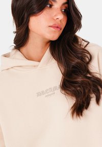 Sudadera con capucha beige hecha de una tela suave, con un estampado tonal de "DREAM". El modelo tiene el cabello castaño ondulado y pecas sutiles.