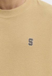Sudadera de punto waffle beige con cuello acanalado y una pequeña letra "S" bordada en negro en el pecho. Textura suave y diseño informal.