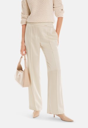 Beige Hosen mit Falten und einer glatten Textur, kombiniert mit einem leichten Strickpullover und einer passenden Handtasche. Spitz zulaufende nudefarbene Absätze vervollständigen den Look.