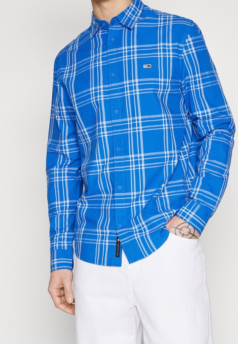 Camicia a scacchi blu con strisce bianche e blu scuro, colletto button-down, maniche lunghe e piccolo logo sul petto. Realizzata in tessuto.
