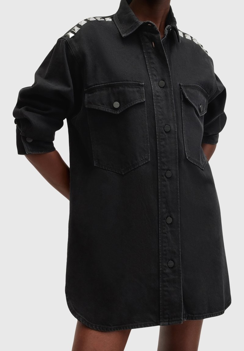 Robe chemise noire surdimensionnée avec manches longues, boutons-pression à l'avant, deux poches à rabat sur la poitrine et clous pyramidaux blancs aux épaules.