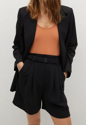 Kvinde iført sort blazer og sorte shorts med høj talje og bælte, kombineret med en ærmeløs orange ribbet top, står med hænderne i lommerne.