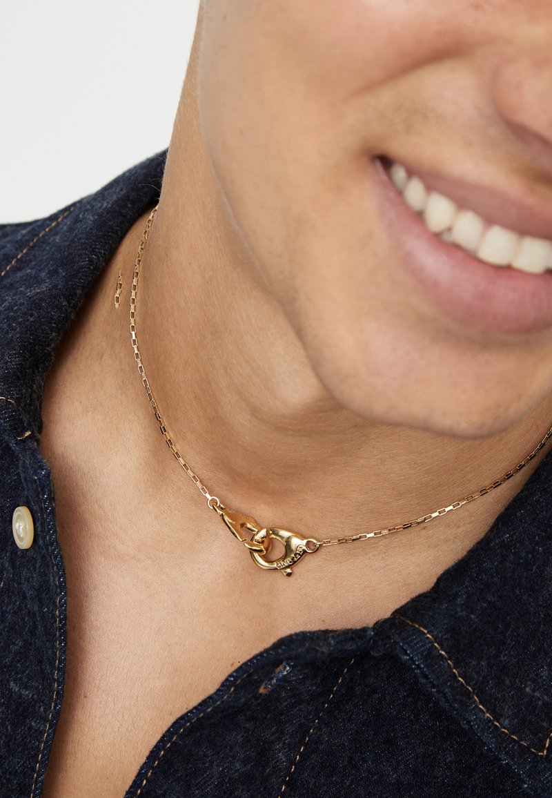 Collar de cadena dorada con diseño de broche y eslabones texturizados. Llevado cerca del cuello, complementando una camisa de mezclilla oscura.