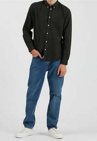 Chemise noire à manches longues avec poche poitrine, associée à un jean bleu clair et des baskets blanches. Fond uni.