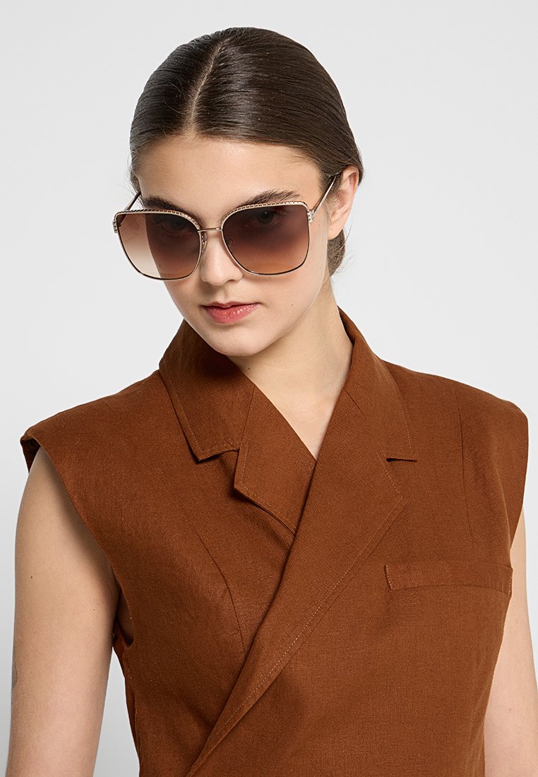 Vrouw met back slick haar, die oversized bruine gradient zonnebril en een mouwloze bruine blazer draagt, tegen een effen grijze achtergrond.