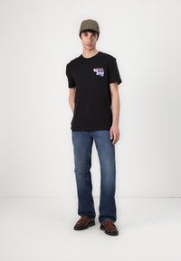 Sort bomulds T-shirt med et pink og blåt grafik på venstre bryst, parret med blå denim jeans og brune sko. Tjekket kasket på hovedet.