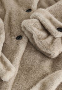 Gesneden zicht op een beige nepbonten jas met een gladde textuur, zwarte knopen en een omgeslagen kraag. Zacht materiaal met een warme uitstraling.