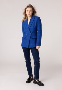 Modrý štruktúrovaný blazer s dvojradovým designom, dvoma prednými vreckami, skombinovaný s tmavými džínami a čiernymi mokasínami.