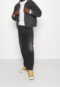 Grå denimjacka över en vit stickad tröja, kombinerad med förbleknade svarta jeans och klara gula sneakers. Minimalistisk design, klassisk passform.