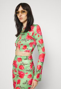 Top crop floral verde com rosas vermelhas, apresentando mangas longas e um detalhe de keyhole na parte frontal; combinado com uma saia lápis estampada a condizer.
