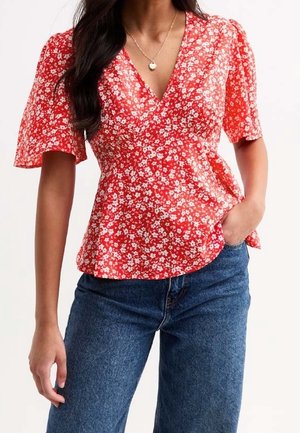 Žena v červené květované halence s výstřihem do V a volnými rukávy, modré džíny, jedna ruka v kapse.