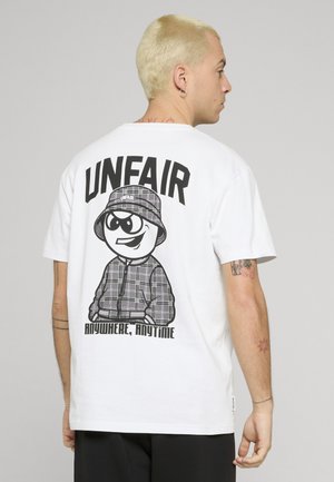 Young man with short blonde hair wearing a white T-shirt featuring a cartoon character in plaid and text "UNFAIR Anywhere, Anytime" on the back.