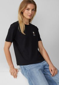 T-shirt en coton noir avec un col rond et des manches courtes, orné d'un petit motif de cupcake brodé sur la poitrine. Porté avec un jean bleu clair.