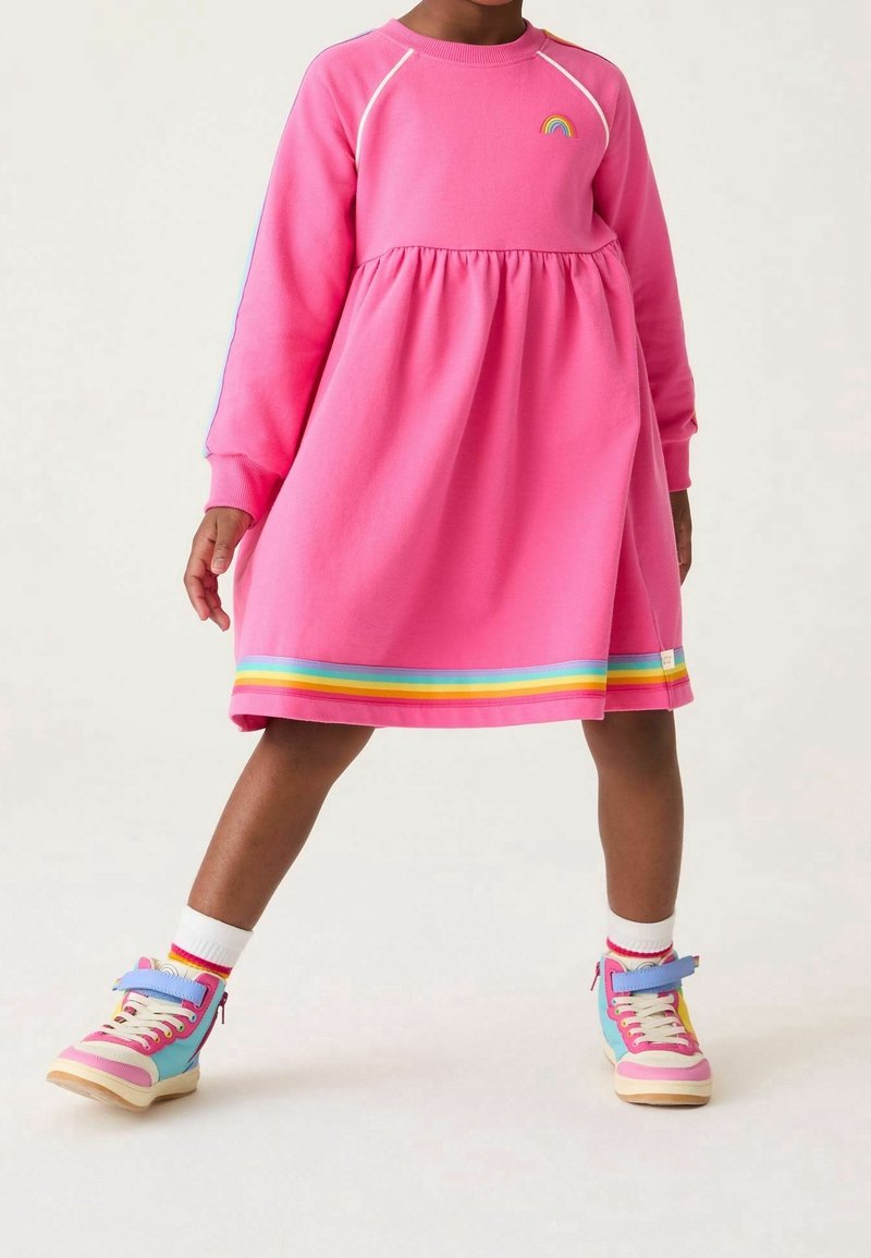 Enfant portant une robe rose à manches longues avec bordure arc-en-ciel et écusson arc-en-ciel, et des baskets montantes colorées avec des chaussettes blanches.