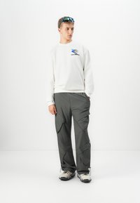 Giovane uomo che indossa una felpa bianca con grafica, pantaloni cargo grigi, scarpe da escursionismo e occhiali da sole sportivi blu sulla testa.