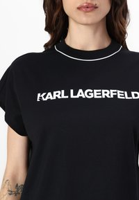 Melna T-kreklis ar baltu "KARL LAGERFELD" logo, noapaļota apkakle ar balto apdari, īsām piedurknēm un mīkstu auduma tekstūru.