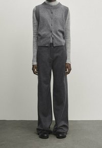 Person wearing a grey buttoned sleeveless cardigan over a light grey long-sleeve top, dark grey wide-leg pants, and black shoes.