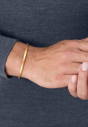 Pulsera de metal con un diseño suave y plano en un brillo dorado, llevada en una muñeca sobre un fondo de suéter gris.