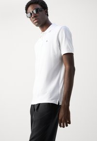 Hvit polotrøye med krage, korte ermer og sidesplitter; kombinert med svarte bukser. Stoffet ser glatt og lett ut.