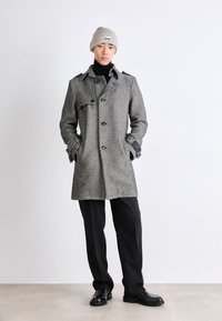 Manteau en chevrons gris avec un design croisé, de grands boutons, des poches latérales et un col cranté, assorti à un pantalon et des chaussures noires.