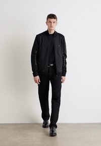 Black zip-up jacket with textured sleeves, worn over a black shirt. Paired with black trousers and black shoes, set against a neutral background.