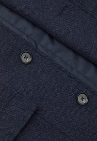 Ciemnoniebieska tkanina o fakturze z gładką podszewką, z czarnym guzikiem z logo. Zawiera kieszeń po lewej stronie.