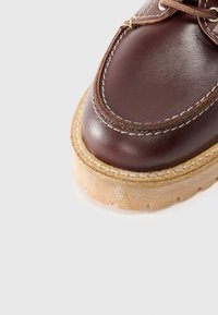 Zapato de cuero marrón con punta redondeada, costuras en contraste y una suela texturizada de color crema. Presenta un diseño de cordones y una construcción robusta.