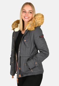 Young woman smiling, wearing a gray winter jacket with a tan faux fur hood and hands in pockets, against a plain background.
