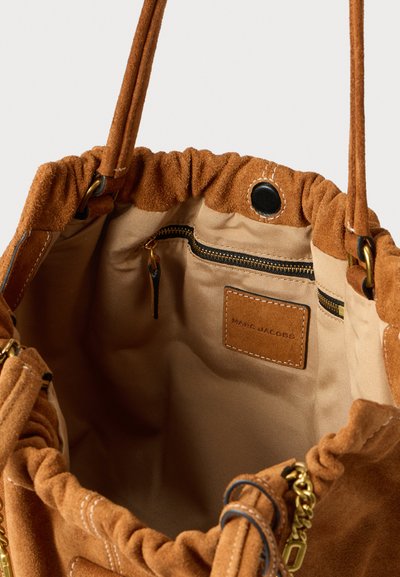 Innenansicht einer braunen Wildlederhandtasche mit gerafftem oberen Design, ausgestattet mit einer Reißverschlusstasche und einem Marken-Lederanhänger in Cognacfarbe. Goldfarbene Beschläge sichtbar.