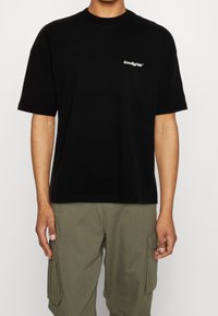 Czarny bawełniany t-shirt z krótkimi rękawami, okrągłym dekoltem i małym białym logo "lowlights" na piersi; zestawiony z zielonymi spodniami typu cargo.