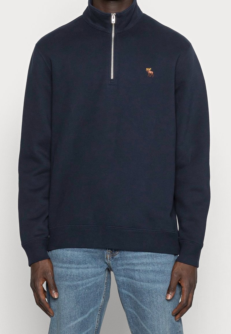Donkerblauwe pullover van zachte stof, met een kraag met een halve rits en een klein geborduurd logo op de borst. Standaard pasvorm met lange mouwen.