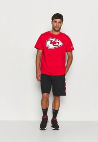 Raudonas marškinėlis su „Kansas City Chiefs“ logotipu, derinamas su juodais šortais. Modelis dėvi raudonus ir juodus sportinius batelius, kulkšnies ilgio kojines, stovi tiesiai.