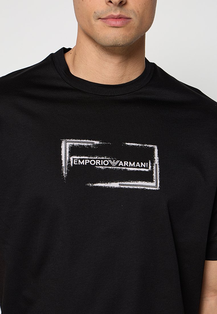 Hombre con camiseta negra con marco rectangular desgastado blanco y logo de texto "EMPORIO ARMANI" centrado en el pecho.