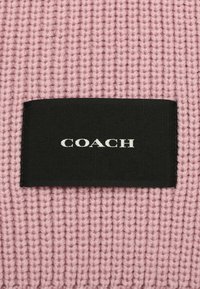 Svetlo roza pletenina z teksturiranim vzorcem, ki vsebuje črno tkaninsko oznako z belo napisano besedo "COACH".