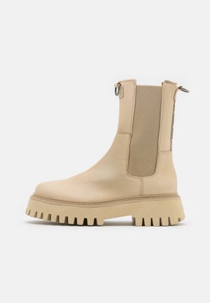 Bottine beige en cuir avec panneaux élastiques, languette arrière et semelle en caoutchouc épaisse à dents. Texture lisse et design minimaliste.