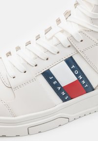 Sneaker bianca con pannelli in pelle e rete, caratterizzata da un patch con marchio blu, rosso e bianco. Lacci bianchi piatta e suola testurizzata.