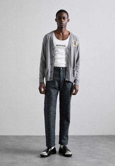 Γκρι ζακέτα πάνω από λευκή μπλούζα, συνδυασμένη με σκούρες παντελόνες με μοτίβο και επωνυμία. Τα παπούτσια είναι μαύρα με γυαλιστερή φινίρισμα.