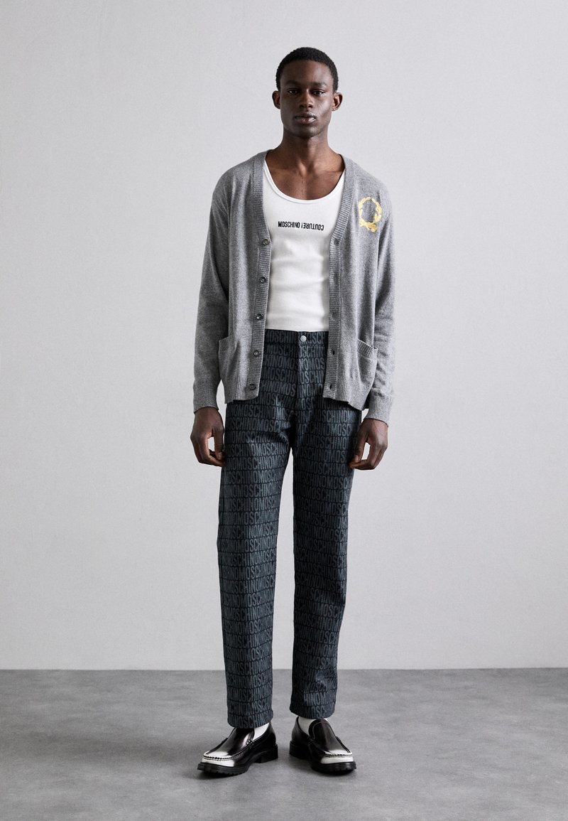 Grau kardigan über einem weißen Tanktop, kombiniert mit dunkel gemusterten Hosen mit einem markierten Druck. Die Schuhe sind schwarz mit einem glänzenden Finish.