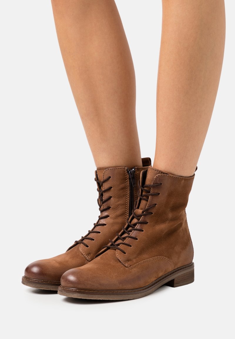 Bottines marron en cuir à lacets avec fermeture éclair latérale portées sur des jambes nues sur un fond uni.
