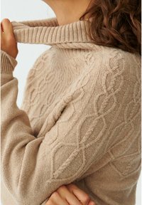 Beżowy sweter z dzianiny z wysokim prążkowanym kołnierzem. Posiada strukturalny wzór w splot kablowy i długie rękawy. Miękka faktura materiału.