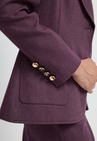 Paarse op maat gemaakte blazer van een gestructureerde stof, met vier gouden knopen op de mouw en een zijzak voor extra detail.