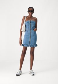 Denim brezramen obleko s centralnim zadrgo, žepi in nabrano obrobo. Oblečena z sivimi supergami in svetlo torbico čez ramo. Črne sončna očala.