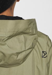 Zielono-oliwkowa kurtka z kapturem o matowej teksturze; ma czarne logo na plecach i czarne podszycie.