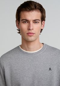 Grauer Sweatshirt mit rundem Halsausschnitt, strukturiertem Stoff und kleinem schwarzen Totenkopf-Logo auf der linken Brust. Klassische Passform und lässiges Design.